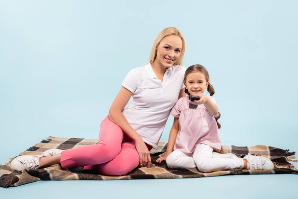 Lächelnde Mutter und Tochter auf Decke sitzend und Film auf blau anschauend - Foto, Bild