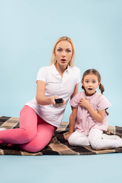 shocked mother and daughter sitting on blanket and watching movie on blue - Foto, Imagen