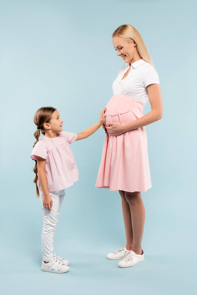 volledige lengte van dochter aanraken buik van zwangere moeder op blauw - Foto, afbeelding