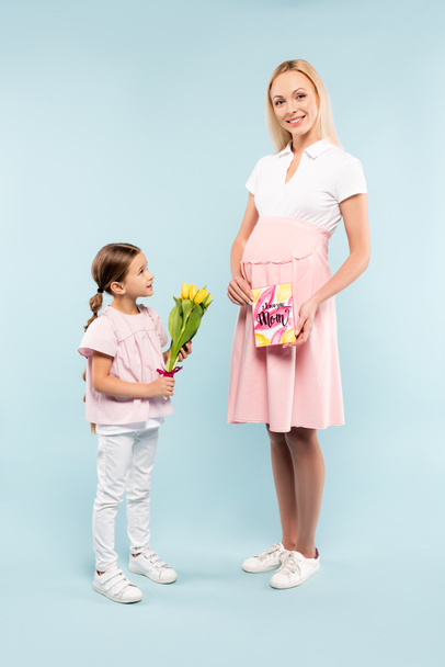 full length of kid holding tulips near happy pregnant mother on blue - Foto, afbeelding