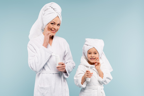 gelukkig moeder en kind in badjassen houden buizen en het aanbrengen van cosmetische crème op gezichten geïsoleerd op blauw - Foto, afbeelding