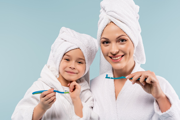 cheerful mother and kid in bathrobes holding toothbrushes with toothpaste isolated on blue - 写真・画像