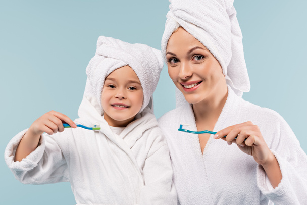szczęśliwa matka i dziecko w szlafrokach trzymających szczoteczki do zębów z pastą wyizolowaną na niebiesko - Zdjęcie, obraz