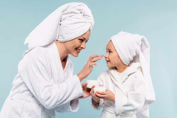happy mother in bathrobe holding container and applying cosmetic cream on face of daughter isolated on blue - Φωτογραφία, εικόνα