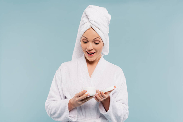 amazed woman in bathrobe holding container with cosmetic cream isolated on blue - Fotoğraf, Görsel