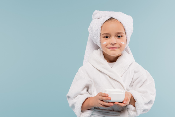 cheerful child in bathrobe and cosmetic cream on face holding container isolated on blue - Photo, image
