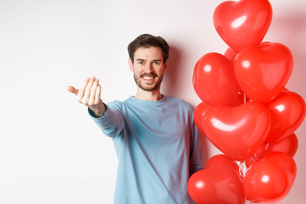 Smiling man beckon you to come closer, follow me gesture, taunting his lover move forward, have romantic surprise, standing near red balloon on valentines day, white background - Photo, Image