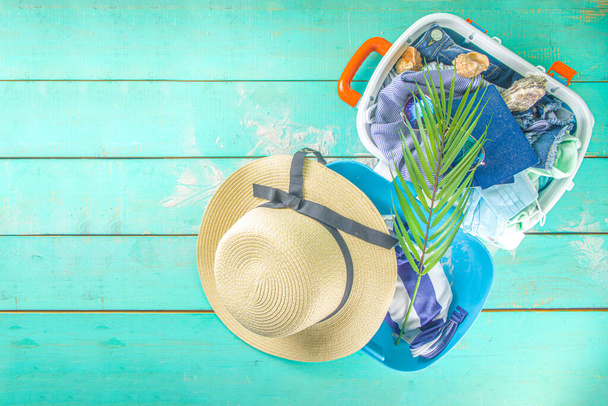 Παραλία και Διακοπές αξεσουάρ για διακοπές σε μπλε σανίδα. Θήκη για ταξίδια, καλοκαιρινά γυναικεία ρούχα, γυαλιά ηλίου, αξεσουάρ και τροπικά φύλλα σε φωτεινό μπλε φόντο - Φωτογραφία, εικόνα
