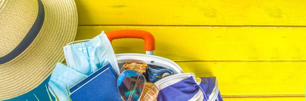 Παραλία και Διακοπές αξεσουάρ για διακοπές σε μπλε σανίδα. Θήκη για ταξίδια, καλοκαιρινά γυναικεία ρούχα, γυαλιά ηλίου, αξεσουάρ και τροπικά φύλλα σε φωτεινό κίτρινο φόντο - Φωτογραφία, εικόνα
