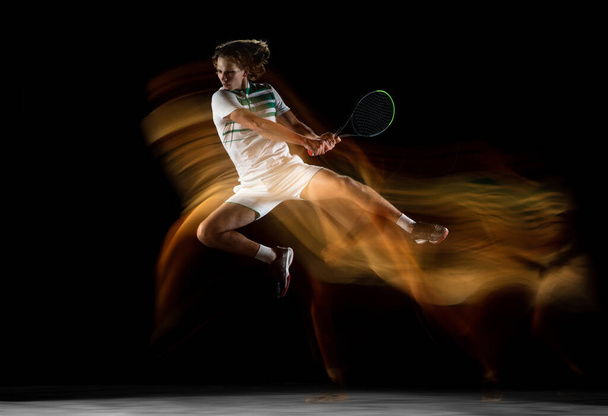 Young caucasian professional sportsman playing tennis on black background in mixed light - Foto, afbeelding