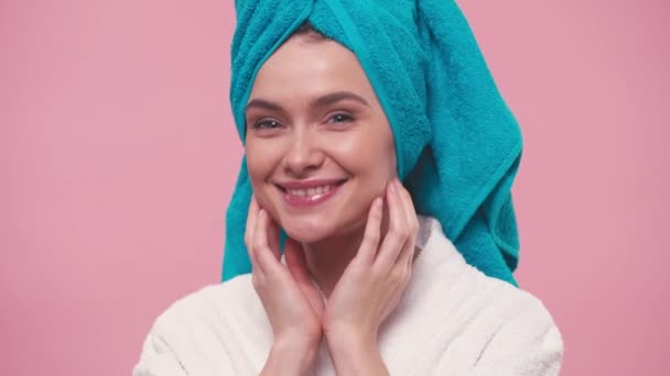 χαρούμενη γυναίκα με πετσέτα στο κεφάλι αγγίζοντας καθαρό πρόσωπο απομονωμένο σε ροζ - Πλάνα, βίντεο