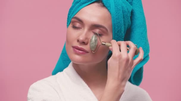 mulher com toalha na cabeça massageando rosto com rolo de jade isolado em rosa - Filmagem, Vídeo