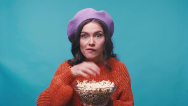 fascinada mujer en boina comiendo palomitas de maíz y viendo películas aisladas en azul - Imágenes, Vídeo