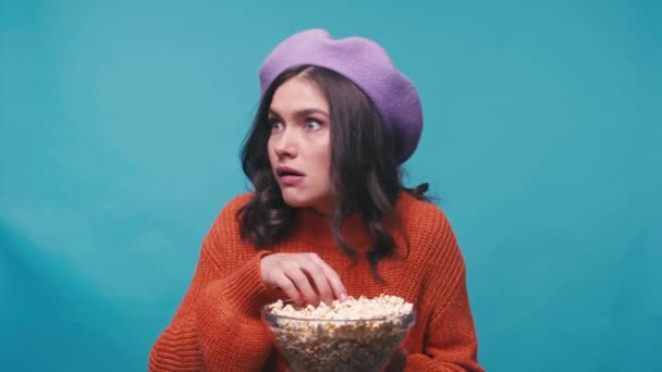 stressed woman in beret holding bowl and eating popcorn on blue - Felvétel, videó