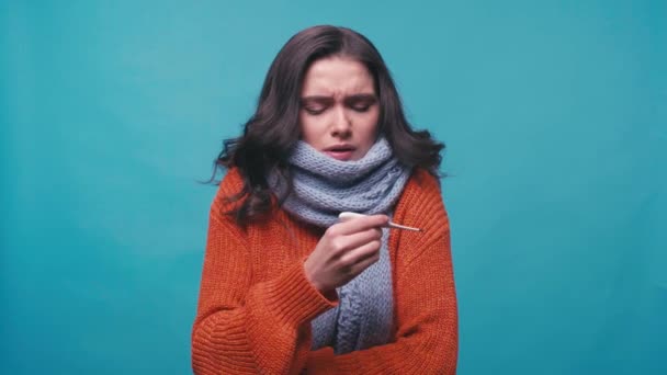 Kranke Frau im Schal blickt auf digitales Thermometer isoliert auf blauem Grund - Filmmaterial, Video