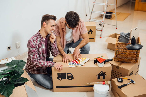 Young gay couple sitting on the floor of a new apartment they moved in, looking at color samples to choose the right color for their wall, while renovating apartment. Moving into a new home. - Foto, Imagen