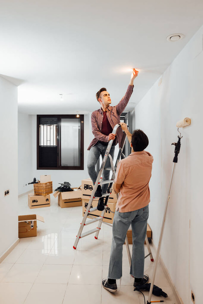 Νεαρός στην κορυφή μιας σκάλας να φτιάχνει μια λάμπα ενώ ο φίλος του βοηθάει να κρατηθεί η σκάλα. Γκέι ζευγάρι κάνει ανακαινίσεις σε ένα νέο σπίτι, διαμέρισμα, με κουτιά από χαρτόνι στο πάτωμα. - Φωτογραφία, εικόνα