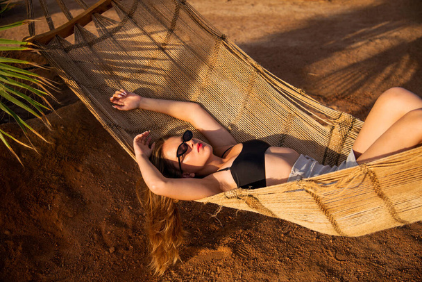 Молода красива дівчина в сонцезахисних окулярах позує лежачи на гамаку, красива жінка відпочиває у відпустці на пляжі, розслабляється, портрет на відкритому повітрі
 - Фото, зображення
