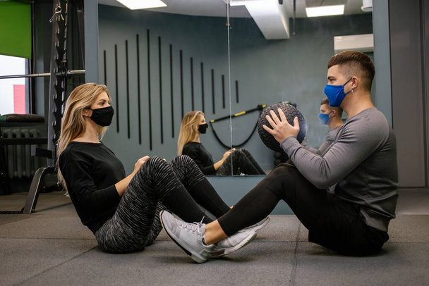 Young woman with protective mask working out with personal trainer at the gym during Covid-19 pandemic. She is pumping up her muscule with dumbbell. Soft focus - Фото, зображення
