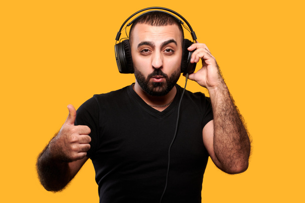 Νεαρός αραβικός dj με γενειάδα που ακούει μουσική στα ακουστικά και δείχνει τον αντίχειρα επάνω στο κίτρινο απομονωμένο φόντο - Φωτογραφία, εικόνα