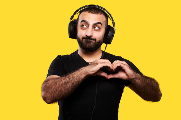 Zabawny pozytywny młody człowiek z brodą i słuchawkami słucha muzyki i pokazuje serce z dłońmi na żółtym, odizolowanym tle. Międzynarodowy Dzień DJ. - Zdjęcie, obraz