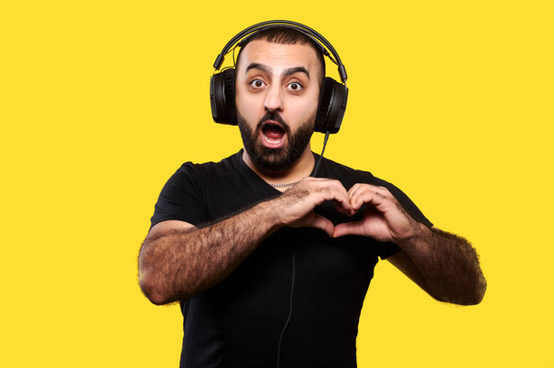 髭とヘッドフォンを持つ面白い正の若い男は音楽を聞き、黄色の孤立した背景に彼の手で心を示しています。国際DJ Day. - 写真・画像