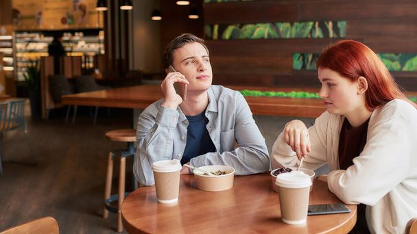Two teenagers spending time together, sitting in a cafe on a daytime. Girl looking bored, eating her meal while her boyfriend is ignoring her, talking on the phone - Фото, изображение