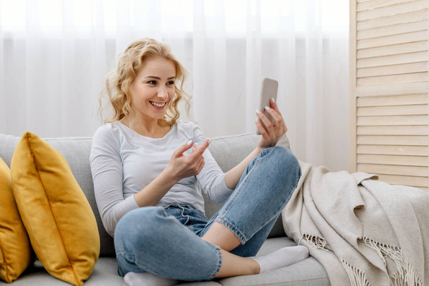 Happy young woman looking at smartphone screen, talking on video call with friends. Smiling female sitting on comfort couch in room, spending morning at home, enjoy weekend day - 写真・画像