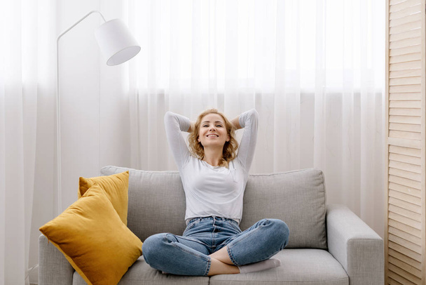 Smiling female freelancer crossing hands behind head, closing eyes, taking a break during remote work day. Happy adult woman in casual clothes on comfort sofa in cozy living room, resting at home - Фото, зображення