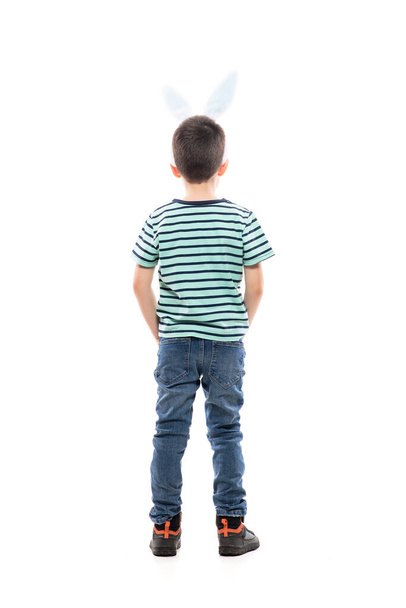 Cooler selbstbewusster kleiner Junge mit Osterhasenohren und Händen in den Hosentaschen, die wegschauen und den Kopierraum betrachten. Volle Länge isoliert auf weißem Hintergrund. - Foto, Bild