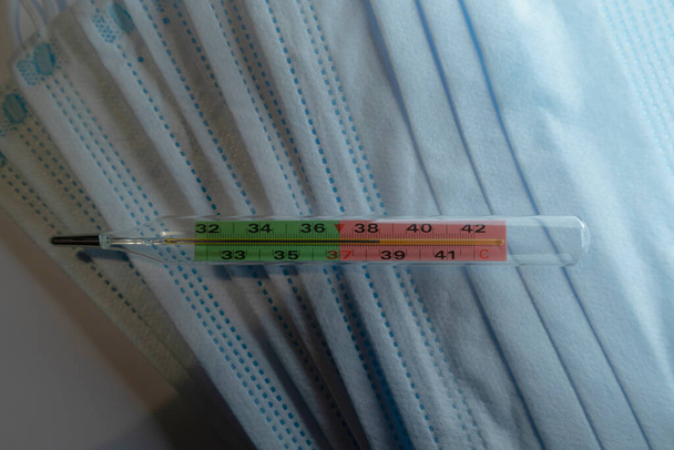 Glass thermometer shows a high temperature. Background of defocused blurred medical masks - Photo, Image