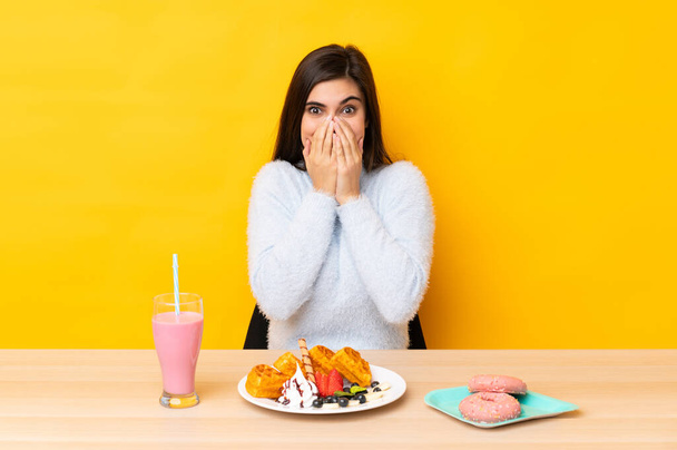 Young woman eating waffles and milkshake in a table over isolated yellow background with surprise facial expression - Fotó, kép