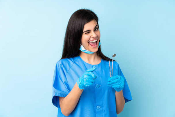Mujer joven dentista sosteniendo herramientas sobre aislado fondo azul señala el dedo en usted - Foto, Imagen