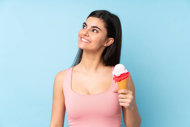 Young woman holding a cornet ice cream over isolated blue background looking up while smiling - Фото, изображение