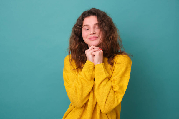Beautiful curly-haired young girl holds her hands under her chin, dressed in fashionable yellow clothes, isolated by a blue background, dreaming with her eyes closed - Fotografie, Obrázek