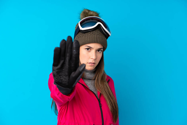 Девушка-лыжница-подросток в очках для сноуборда на изолированном синем фоне делает жест остановки рукой - Фото, изображение
