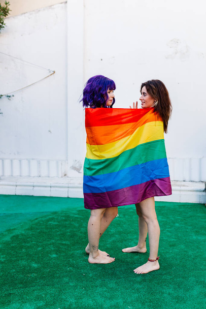 Arka bahçesinde gökkuşağı bayrağı olan iki kadın. Gökkuşağı bayrağı tutan ve mutlu bir şekilde kucaklaşan birkaç lezbiyen kız. Bir çift lezbiyen kadın. - Fotoğraf, Görsel