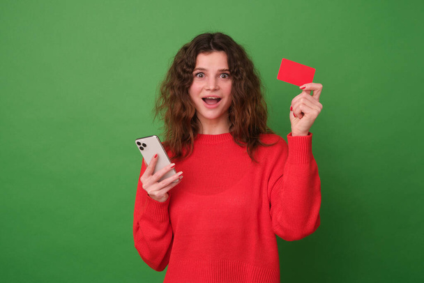 A beautiful girl in a red sweater on a green background. The girl in a surprised mood is holding a red credit card in her hand and a phone in her other hand, her gaze is directed at the camera - Fotografie, Obrázek