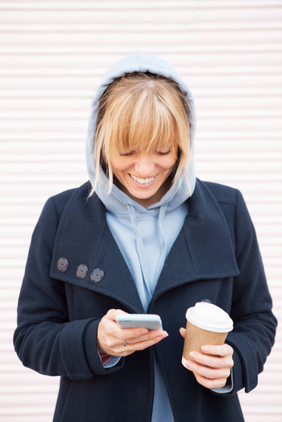 Cabello rubio mujer joven con capucha azul está utilizando el teléfono inteligente y beber tomar tomar café mientras camina al aire libre en la ciudad. Expresión de la cara feliz, sonriendo con dientes.  - Foto, Imagen