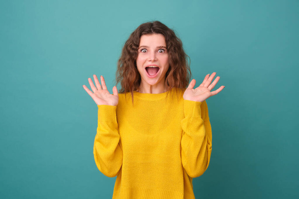 Amused cute girl with curly hair, raises her palms, has a cheerful expression, smiles widely, sees something funny, wears a yellow sweater isolated on a blue background - Photo, image