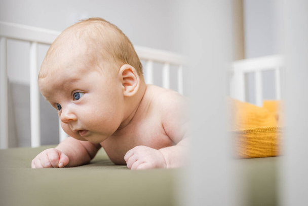 Buclatý novorozenec, zabalený do módní žluté mušelínové přikrývky, se pokouší zvednout hlavu, zatímco leží na břiše v bílé dřevěné postýlce. Koncept zdravého vývoje miminka 0-6 měsíců, péče o miminko.  - Fotografie, Obrázek