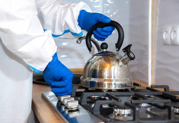 Doctor wearing medical protective suit, goggle, mask, and gloves preparing tea on the kitchen. Teapot on the gas stove. Protection mers by virus epidemic. Coronavirus (COVID-19). Healthcare concept. - 写真・画像