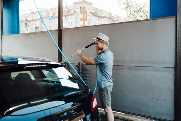 Young man with hat washing his car during daylight at car wash station using high pressure water. - Φωτογραφία, εικόνα