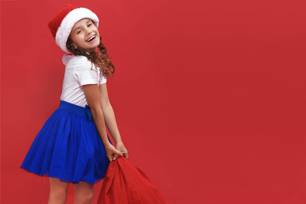 Entzückende kleine lächelnde Mädchen mit Weihnachtsmütze tragen Geschenktüte isoliert auf rotem Hintergrund. Raum für Text. - Foto, Bild