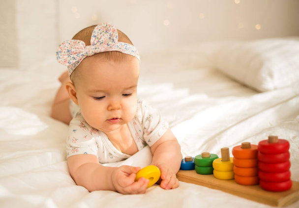 Κοριτσάκι βρίσκεται σε ένα χρωματιστό κοστούμι σώματος και μια κορδέλα και παίζει με ένα εκπαιδευτικό παιχνίδι σε μια λευκή κουβέρτα στο κρεβάτι. Πρόωρη ανάπτυξη παιδιών - Φωτογραφία, εικόνα