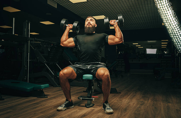 Muscular young man lifting weights in a dark gym - Φωτογραφία, εικόνα