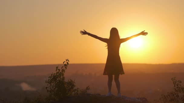 Молода жінка в літньому платті стрибає на відкритому повітрі з витягнутими руками насолоджуючись видом на яскраво-жовтий захід сонця
. - Кадри, відео
