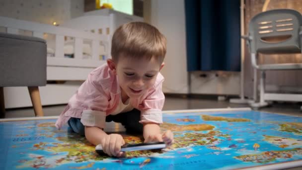 Kleine lachende jongen liggend op de vloer in de slaapkamer en kijkend op de grote wereldkaart door vergrootglas. Concept van reizen, toerisme en onderwijs voor kinderen. Verkenning en ontdekkingen van kinderen. - Video