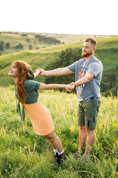 young beautiful couple red-haired girl in a pink dress and green jacket a man in a gray t-shirt and green shorts are having fun in the grass in a field in nature at sunset - Fotografie, Obrázek