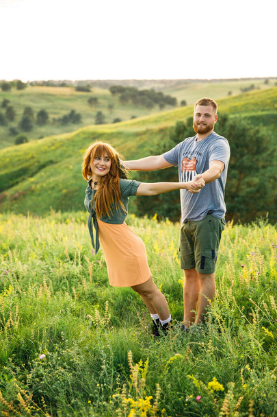 young beautiful couple red-haired girl in a pink dress and green jacket a man in a gray t-shirt and green shorts are having fun in the grass in a field in nature at sunset - Foto, Imagem